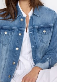 Vaaleansininen farkkutakki, jossa napitus edessä, kaksi rintataskua ja kulutettuja yksityiskohtia. Varustettu metallinappien ja muotoillun kauluksen kanssa.