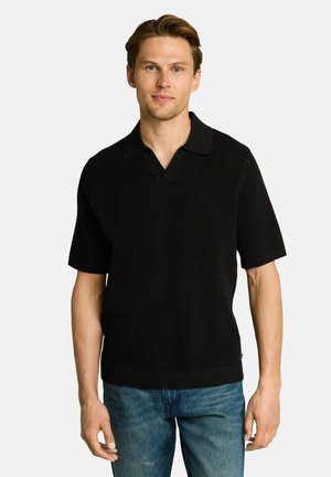 Young man with light brown hair wearing a black short-sleeve knit polo shirt and blue jeans standing against a plain background.