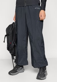 Črne lahke hlače Terrex z gladko teksturo, elastičnim pasom in zadrgami. V kombinaciji s črnimi pohodniškimi čevlji in nahrbtnikom.