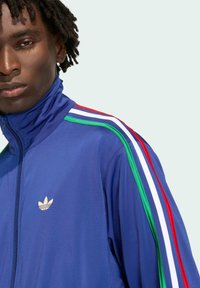 Blue zip-up track jacket featuring a high collar and three contrasting stripes in red, green, and white along the shoulders. Gold logo present.