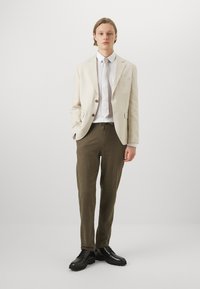 Linen blazer in beige with two buttons, paired with olive green relaxed-fit pants and black lace-up shoes, set against a neutral backdrop.