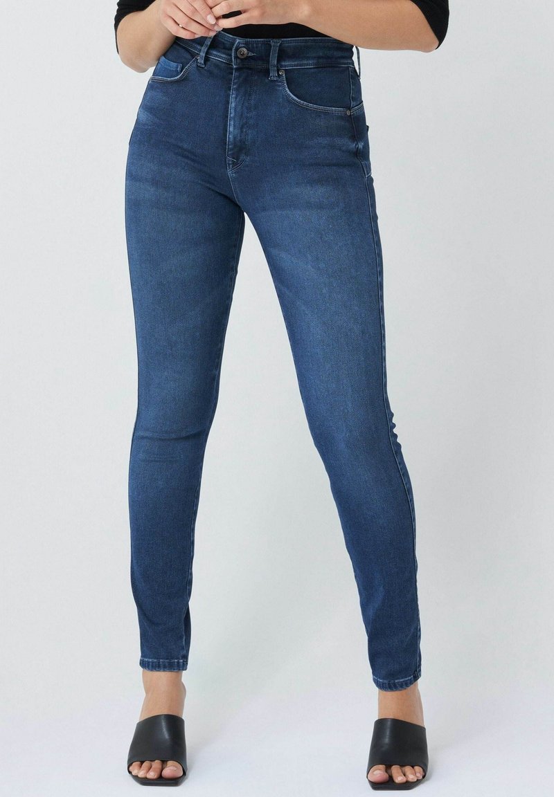 Femme portant un jean skinny taille haute bleu foncé et des chaussures noires ouvertes à bouts ouverts, debout devant un fond clair uni.