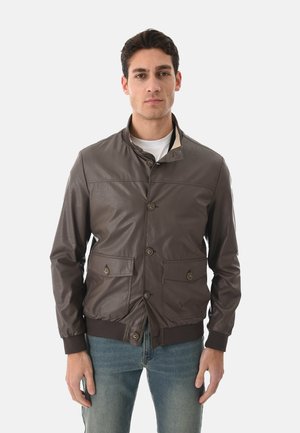 Uomo con giacca di pelle marrone abbottonata con due tasche frontali a bottone e jeans blu, in piedi contro uno sfondo chiaro uniforme.