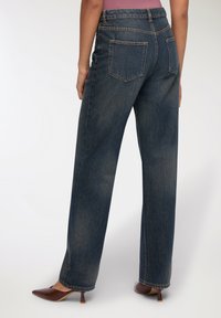 Jeans in denim scuro a gamba larga con vita alta, due tasche posteriori e dettagli di cucitura visibili, abbinati a mule con tacco marroni.