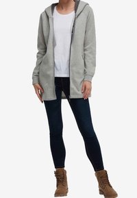 Sudadera gris con cremallera y capucha ajustable, llevada sobre una camiseta blanca, combinada con jeans ajustados de color azul oscuro y botas marrones con cordones.