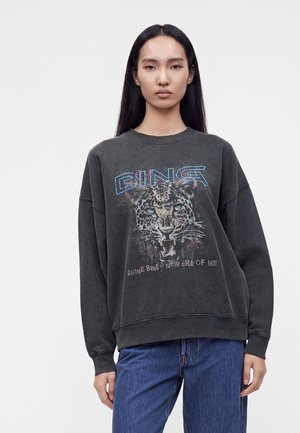 Jeune femme aux longs cheveux foncés porte un sweat-shirt noir délavé montrant un léopard qui grogne et le texte "BING" et "A new era of me."