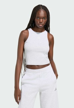 Młoda kobieta z długimi warkoczami ubrana w jasnoszary bezrękawnik typu crop top oraz dopasowane spodnie dresowe, z rękami w kieszeniach.