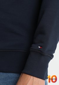 Primer plano de sudadera azul marino con puños acanalados, costuras planas y un pequeño detalle de logo en rojo, blanco y azul en la manga.