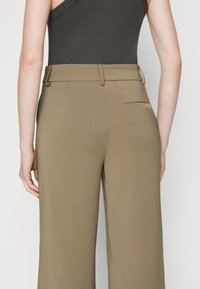 Vidbenade, ribbad byxor i khaki, med en slät textur, skräddarsydd midja och en bakficka på höger sida.