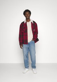 Camicia di flanella a quadri rossi e neri con colletto in pelliccia bianca, indossata sopra una maglietta beige e jeans blu, abbinata a sneaker bianche.