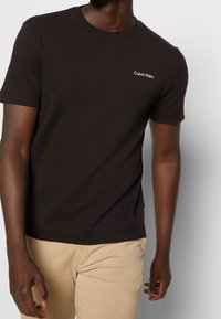 Černé bavlněné tričko s krátkým rukávem, které má malé bílé logo "Calvin Klein" na levé straně hrudi. Jednoduchý design s kulatým výstřihem.