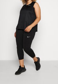 Černý atletický top a capri legíny vyrobené z prodyšného materiálu, s bílým logem Nike na obou kusech. Černé tenisky doplňují outfit.