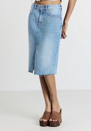 Gonna midi in denim blu chiaro con spacco frontale indossata con sandali con tacco a piattaforma marroni, su una persona in piedi contro uno sfondo semplice.