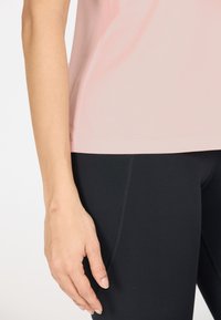 Vaaleanpunainen nopeasti kuivuva paita, joka on yhdistetty mustiin legginsseihin. Asusteessa on sileä rakenne ja tiukka muotoilu. Asu korostaa mukavuutta ja toiminnallisuutta.