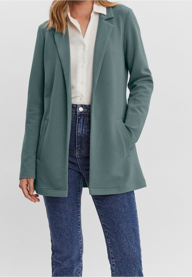 Femme portant un blazer côtelé turquoise par-dessus une chemise blanche à boutons et un jean bleu taille haute, les mains dans les poches du blazer.