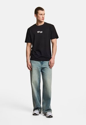 Młody mężczyzna stoi, ubrany w czarną koszulkę z nadrukiem, jasne szerokie dżinsy i białe trampki, patrzy w lewo.