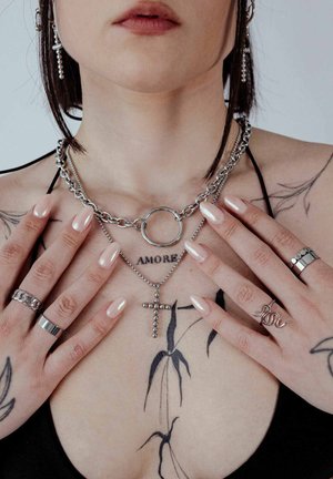 Silberne Kettenhalsketten, eine mit einem Kreisanhänger und die andere mit einem Kreuz, kombiniert mit verschiedenen silbernen Ringen. Leicht lackierte Nägel und sichtbare Tattoos.