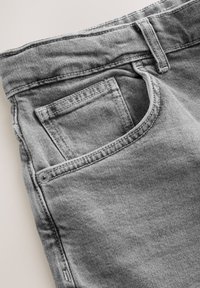 Primo piano di jeans in denim grigio chiaro che mostra la tasca frontale con taschino porta monete, la cintura e i dettagli delle passanti per cintura.