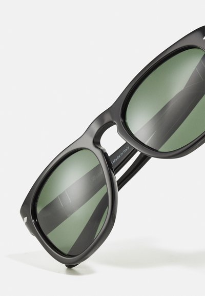Black sunglasses with green lenses, featuring a thick frame and a rounded shape. Note the engraved logo on the inside temple.