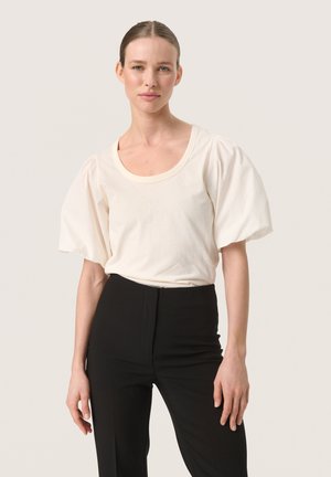 Cremefarvet bomulds t-shirt med bred rund halsudskæring og puf korte ærmer, parret med højtaljede sorte bukser.