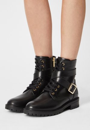 Bottes cheville en cuir noir avec une sangle à boucle, des œillets dorés et une semelle en caoutchouc texturée. Dotées de lacets et d'une fermeture éclair intérieure.