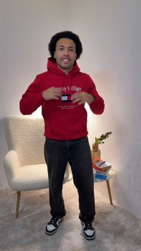 Rotes Hoodie mit "Tommy Hilfiger"-Logo und Farbblock-Design; schwarze Jeans; schwarz-weiße Turnschuhe; Innenraum mit einem Stuhl und Büchern.