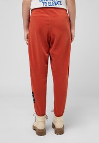 Roodbruin fleece joggers met elastische manchetten en een patch op het rechterbeen, gecombineerd met crèmekleurige veterschoenen op een grijze achtergrond.