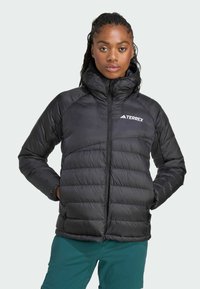 Chaqueta acolchada negra con cuello alto y cierre de cremallera, que presenta un diseño acolchado y logo en el lado izquierdo del pecho. Textura suave y aislante.