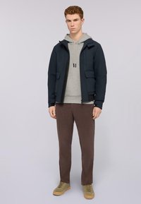 Giacca navy con zip, dotata di due tasche frontali, sovrapposta a un felpa grigia. Pantaloni marroni e sneaker beige completano l'outfit.