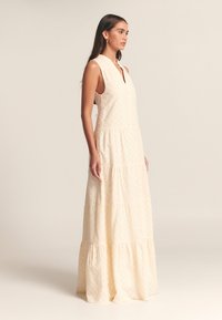 Svetlo bež maxi obleka z teksturiranim vzorcem, brez rokavov, zapenjanjem na gumbe in večstopenjskim robom. Gladek material, sproščena silhueta.