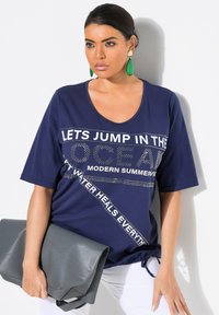 Navyblauwe T-shirt met korte mouwen, ronde hals en bedrukt tekst. Inclusief een knoopdetail aan de zijkant. Grijze tas die in de linkerhand wordt vastgehouden.