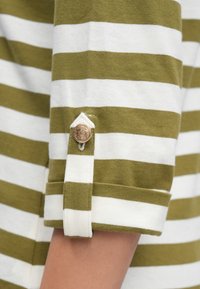 Zaļi un balti svītraina krekla ar sarullētām piedurknēm, kas rotāts ar dekoratīvu pogu un nelielu auduma cilpu netālu no pogas. Kokvilnas materiāls.