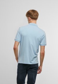 Gaiši zils kokvilnas polo krekls ar apkakli un īsām piedurknēm. Aizmugures skats rāda gludu audumu bez redzamām rakstām.