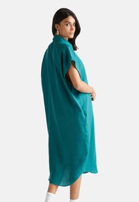 Turkusowa sukienka z lnu o luźnym kroju, z krótkimi rękawami i kołnierzykiem. Hem jest lekko zaokrąglony, a po bokach widoczne są kieszenie.