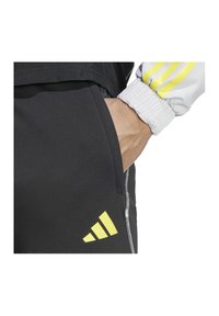 Zwarte sportbroek met een zijzak, voorzien van een opvallend gele Adidas-logo en een gestructureerde stof. De tailleband is elastisch.