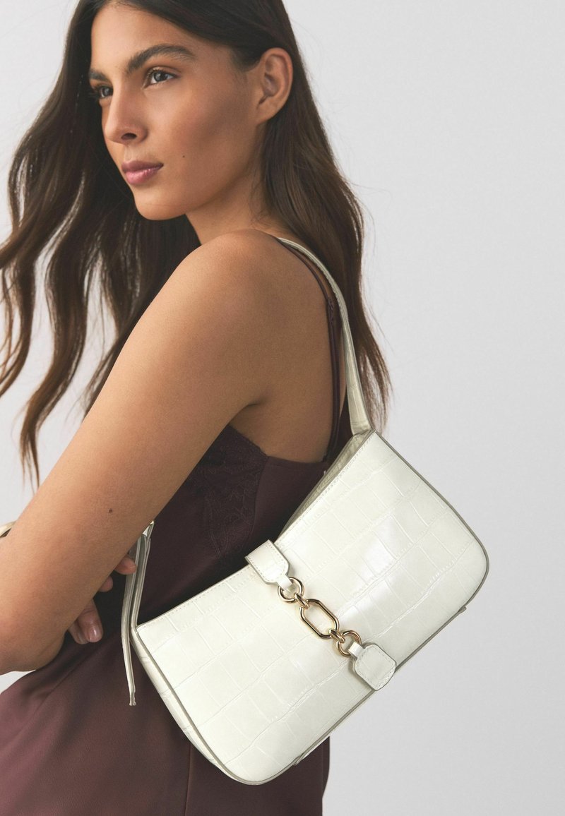 Donna con canotta marrone senza maniche che porta una borsa a tracolla bianca con dettagli in catena dorata su uno sfondo uniforme.