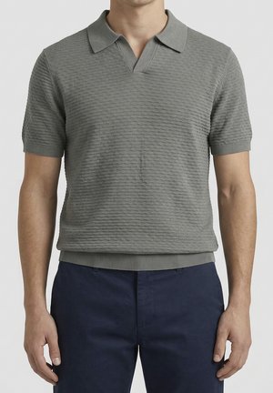 Čovjek odjeven u kratkih rukava teksturiranu sivu polo majicu s rebrastom kragnom i tamnoplave hlače, stoji ispred jednobojne pozadine.