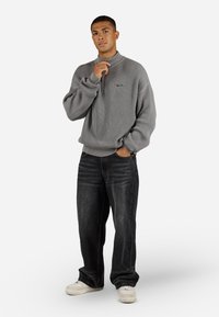 Grijze ribgebreide pullover met een hoge kraag en een voorkantzak, gecombineerd met baggy donkere spijkerbroek en lichte sneakers. Bevat geborduurd logodetail.