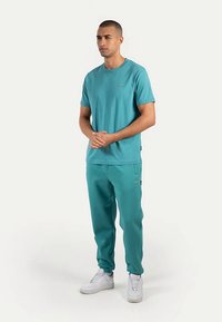 Turkis bomuld t-shirt og matchende joggingbukser, begge med en glat tekstur; har lille logo og sidelommer; hvide sneakers med snørebånd.