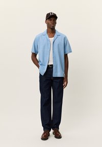 Ανοιχτό μπλε κοντομάνικο πουκάμισο με κουμπιά, λευκό εσώρουχο, σκούρο μπλε τζιν σε χαλαρή εφαρμογή, καφέ δερμάτινα παπούτσια και σκούφο σκούρου χρώματος με λευκές λεπτομέρειες.