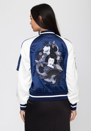 Kobieta nosząca granatową i białą kurtkę bomber z haftowanym japońskim motywem gejszy, kota, karpia koi, wachlarzy i żurawia na plecach.