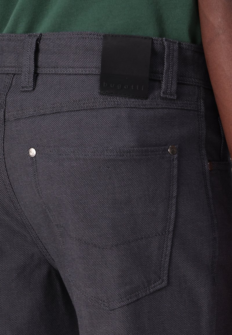 Donkergrijze denimbroek met een gestructureerde afwerking, voorzien van een zwart lederen label met de tekst "bugatti" en zilveren hardware op de zakken.