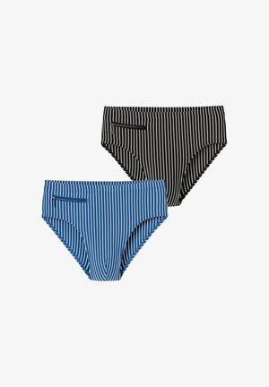 Jeune homme ajustant un slip de bain noir à rayures verticales blanches, debout devant un fond clair uni.