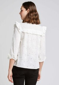 Blouse blanche brodée avec un col volanté, des manches trois-quarts à revers et un motif découpé floral.