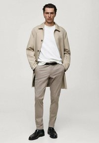 Vaaleanbeige trenchcoat valkoisen t-paidan päällä, yhdistettynä vaaleanbeigeihin housuihin ja mustiin nahkakenkiin. Yksinkertainen muotoilu nappiyksityiskohdilla.