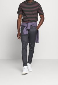 T-shirt rayé vert foncé avec des lignes rouges, pantalon en denim gris et baskets blanches. Sweat-shirt violet noué autour de la taille.