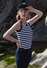 Pasiasty top bez rękawów w czarno-białe paski, w połączeniu z ciemnoniebieskimi dżinsami. Modelka w czarnej czapce, pozująca na tle skalistym.