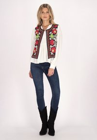 Färgglad broderad väst med blommönster, bärs över en vit blus, i kombination med mörka jeans och svarta cowboyboots.