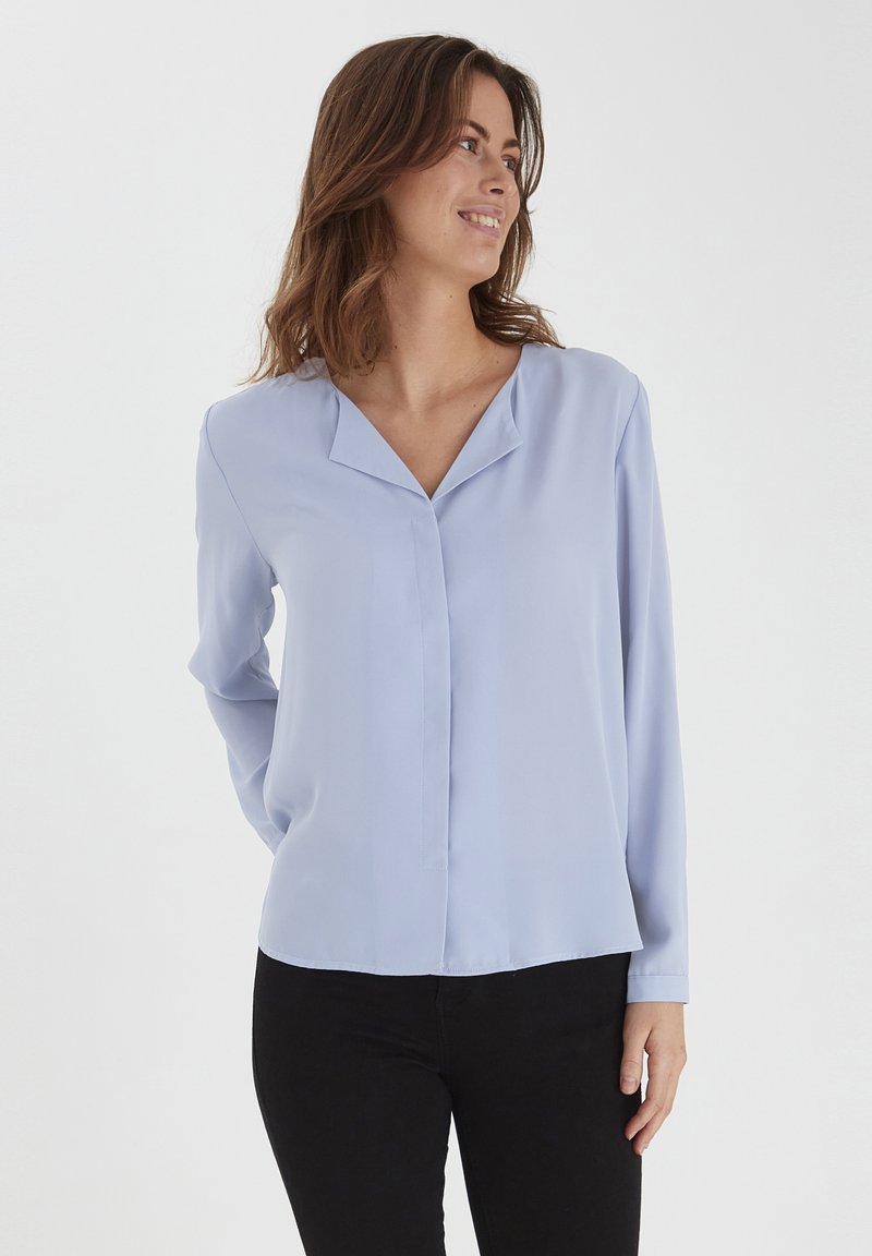 Blusa azzurra chiara con scollatura a V, maniche lunghe e una texture liscia e fluida. Presenta una cucitura frontale e un orlo leggermente curvo.