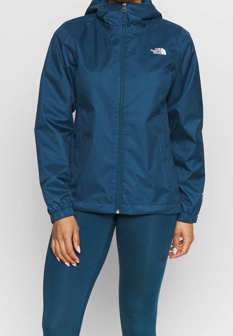 Personne portant une veste à capuche zippée bleue et un legging assorti avec le logo The North Face sur la poitrine de la veste et sur la cuisse du legging.